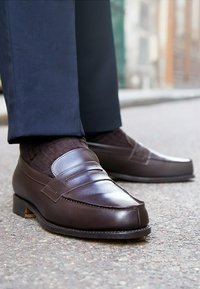 Bruine leren loafers met een gladde afwerking, verhoogde stiknaad en een lage gestapelde hak, gecombineerd met donkere gestructureerde sokken en een broek.