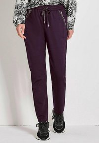 Paarse joggingsbroek met elastische tailleband, koord en ritszakken. Glad stofstructuur, tapered pijpdesign, gedragen met zwarte sneakers.