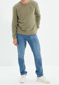 Maglione verde oliva con collo rotondo e stampa grafica sul lato sinistro del petto, abbinato a jeans in denim blu e scarpe da ginnastica bianche.