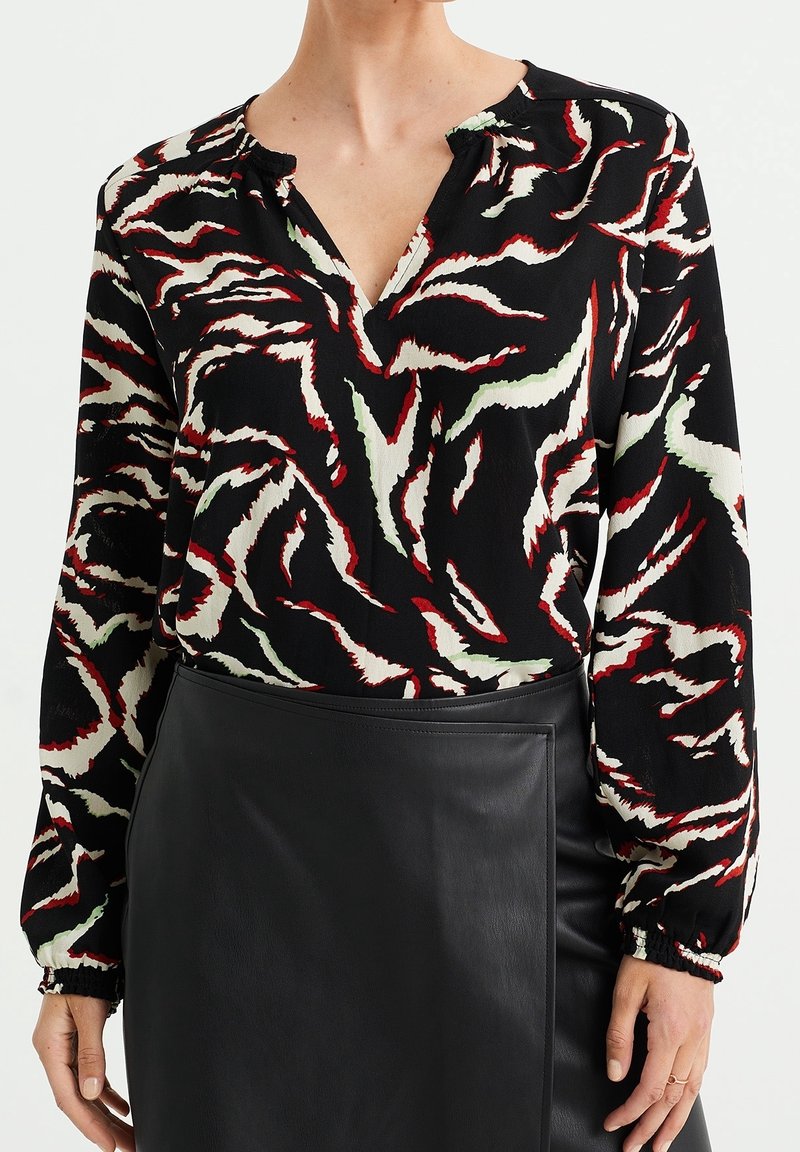 Femme portant un chemisier à motifs abstraits noirs, rouges et blancs à manches longues, rentré dans une jupe en cuir noire.