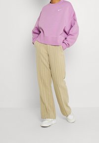 Person som bär en lilag Nike-hoodie, beige randiga byxor, vita sneakers, står med händerna i fickorna mot en helt vit bakgrund.