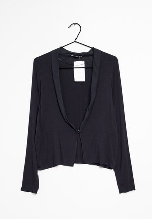 Cardigan nero a maniche lunghe con rever in satin, appeso a una gruccia nera su sfondo bianco, etichetta del negozio attaccata all'interno del colletto.