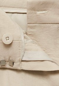 Tissu beige avec une texture lisse, présentant une fermeture à bouton et une quincaillerie en métal. Des coutures apparentes et un détail de poche cachée sont visibles.