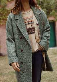 Green herringbone wool overcoat with large buttons, layered over a patterned sweater. Model wears dark jeans. Background features blurred greenery.