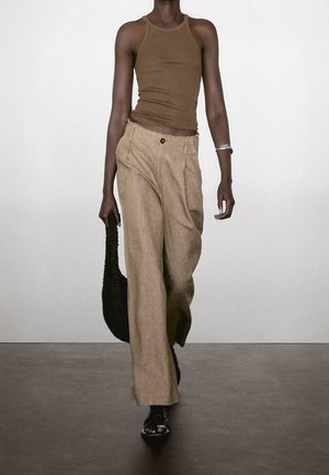 Person walking wearing a brown sleeveless top, loose beige pants, black sandals, silver bracelet, and holding a black woven bag.