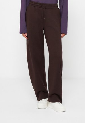 Personne portant un pantalon large marron foncé avec taille à cordon, un haut violet à manches longues et des baskets blanches, debout sur un sol blanc.