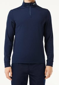 Pull-over bleu marine à manches longues avec col zippé, tissu texturé et logo Lacoste vert sur le col.
