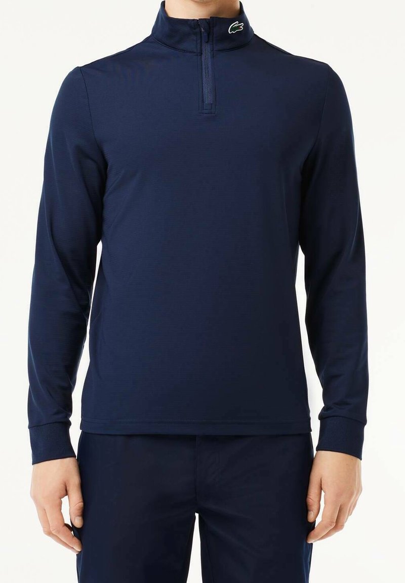 Pull-over bleu marine à manches longues avec col zippé, tissu texturé et logo Lacoste vert sur le col.