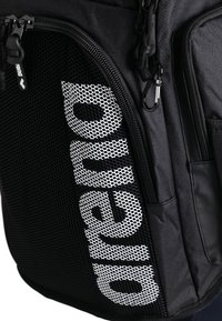 Sac à dos noir avec une poche en maille portant un texte blanc, des zips robustes et un mousqueton. Construit à partir d'un tissu durable au toucher lisse.
