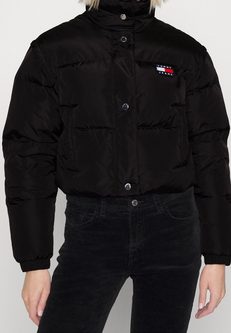 Sort pufferjakke med en kort model, der har en høj krave, trykknapper og en lille logopatch. Materialet har tekstur og varm isolering.