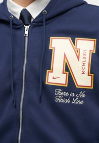 Artimiausias vaizdas į tamsiai mėlynį užtrauktuku užsegamą džemperį su dideliu „N“ logotipu, romėniškais skaitmenimis MCM LXXII ir užrašu „Ten nėra finišo linijos.“