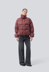 Frau in einer bordeauxroten Daunenjacke mit hohem Kragen und Zugbändern an den Ärmeln, kombiniert mit weit geschnittenen schwarzen Jeans und schwarzen Schuhen, stehend vor grauem Hintergrund.