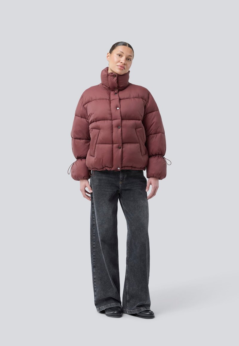 Frau in einer bordeauxroten Daunenjacke mit hohem Kragen und Zugbändern an den Ärmeln, kombiniert mit weit geschnittenen schwarzen Jeans und schwarzen Schuhen, stehend vor grauem Hintergrund.