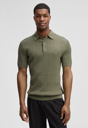 Mann trägt ein olivgrünes, kurzärmeliges Poloshirt mit Zopfmuster und schwarze Hose, steht vor einem einfarbigen hellgrauen Hintergrund.