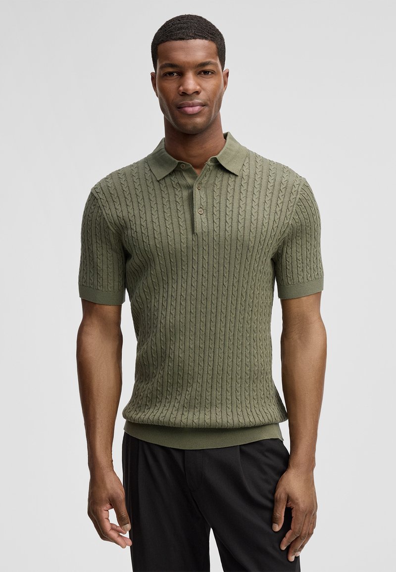 Man die een olijfgroen, kabelgebreid poloshirt met korte mouwen en een zwarte broek draagt, staand tegen een effen lichtgrijze achtergrond.