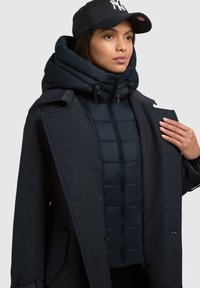 Cappotto esterno blu navy con colletto alto, sovrapposto a un gilet interno imbottito. Presenta una chiusura con zip e cappuccio, con un berretto nero. Texture liscia.