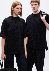 Μαύρα μπλουζάκια από μαλακό ύφασμα με ανάγλυφο σχέδιο. Χαλαρή εφαρμογή, στρογγυλή λαιμόκοψη και χαρακτηριστικές λογοτύπικές λεπτομέρειες.