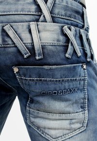 Jeans en denim dans un délavé moyen avec une texture usée, comportant plusieurs passants de ceinture, des accents cousus et un logo sur la poche arrière.