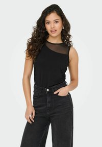 Top noir sans manches avec un panneau transparent sur une épaule, associé à un jean noir taille haute. Texture lisse et design ajusté.