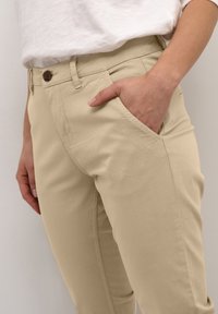 Person som bär beige byxor med en hand vilande i den främre fickan, tillsammans med en vit topp instoppad.