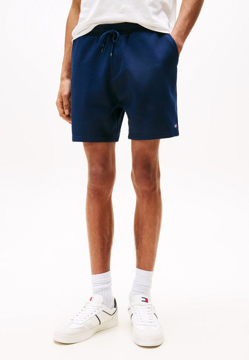Navy-blaue Baumwoll-Shorts mit elastischem Bund und Kordelzug. Weiße Sneakers mit schwarzen Akzenten und weißen Socken vervollständigen das Outfit.