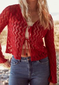 Cardigan rouge en maille présentant un motif en zigzag, des manches longues et une fermeture devant nouée. Porté sur un haut clair et associé à un jean bleu.