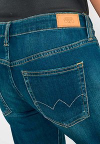 Jean en denim bleu foncé avec des coutures orange, présentant une étiquette en cuir au niveau de la taille et un design de poche unique avec un motif en vagues.