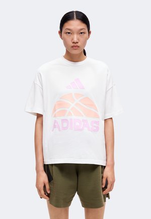 Młoda osoba z ciemnymi włosami w białej koszulce Adidas z różową grafiką koszykówki oraz oliwkowo-zielonych szortach, stojąca na jasnym tle.