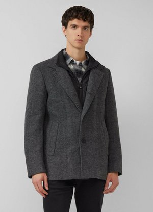 Młody mężczyzna ubrany w ciemnoszary wełniany płaszcz na czarnej pikowanej kurtce i koszuli w kratę, stojący na tle gładkiego jasnoszarego tła.