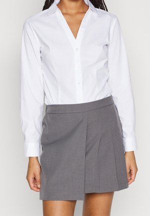 Vrouw draagt een wit overhemd met knopen en een grijze geplooide rok, staand tegen een vlakke lichte achtergrond.
