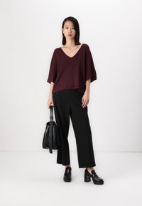Bordeaux gebreid V-hals sweater met korte mouwen, gecombineerd met zwarte wijde broek en zwarte plateau instappers, met een zwarte tas in de hand.