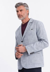 Hellgrauer Leinenblazer mit strukturiertem Finish, einem Ein-Knopf-Verschluss und zwei vorderen Taschen. Verfügt über dunkle Knöpfe und einen Reverskragen.