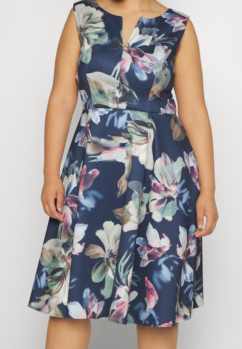 Robe bleu marine sans manches avec de grandes impressions florales pastel et un léger col en V, portée par une personne montrée du cou aux genoux.