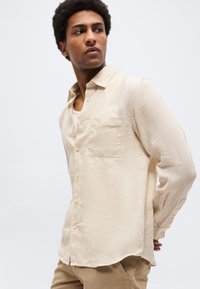 Άνδρας με μπεζ πουκάμισο λινό με κουμπιά και καφέ παντελόνι, κοιτάζοντας πλάγια σε απλό φωτεινό φόντο.