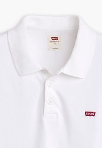 Biała koszulka polo z fakturowanej tkaniny, z kołnierzykiem, z trzema guzikami i małym czerwonym logo Levi's na lewej piersi.