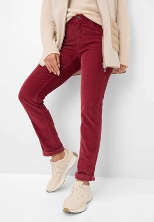 Pantaloni in corduroy rosso con una vestibilità slim e orli arrotolati, abbinati a sneakers beige chiare. Il tessuto ha una texture a coste.