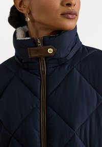 Námornícky prešívaný kabát s vysokým golierom a hnedým koženým pásikom so zlatým gombíkom. Viditeľný zlatý zips a mäkká vnútorná podšívka.