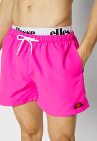 Shorts de bain roses vifs avec une taille élastique, un cordon blanc et des poches latérales. Ils présentent un petit logo multicolore en bas.