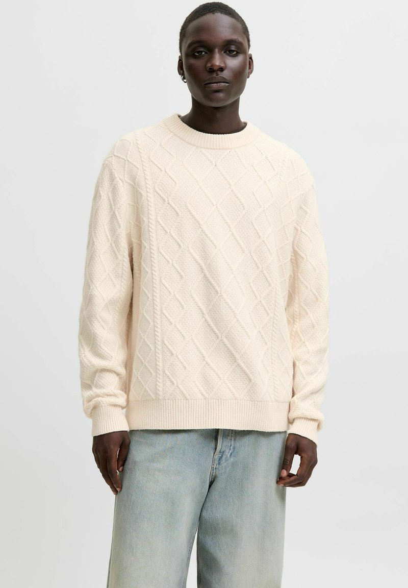 Cremfarbener gestrickter Pullover mit einem strukturierten Rautenmuster, geripptem Ausschnitt und Bündchen. Kombiniert mit hellblauen Schlagjeans.