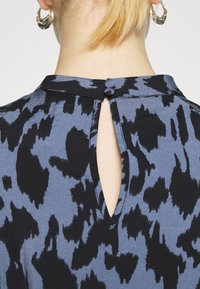 Haut bleu et noir à motif, avec un détail dos en forme de trou rond, tissu doux et fermeture par bouton au niveau de l'encolure.