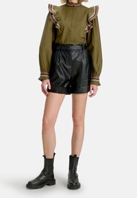Blusa verde oliva con maniche volanti e dettagli rosa, abbinata a pantaloni corti in pelle sintetica nera e stivali neri alti fino al ginocchio.