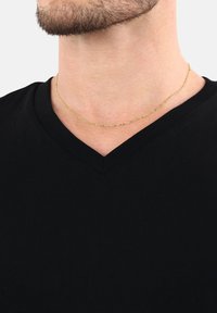 Primer plano de un hombre que lleva una delgada cadena de oro y una camiseta negra de cuello en V contra un fondo liso.