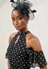 Crna haljina s točkicama, dekolteom u obliku ključa i off-shoulder krojem s blago raširenim rukavima. Uključuje prozirni crni fascinator s mašnicama.