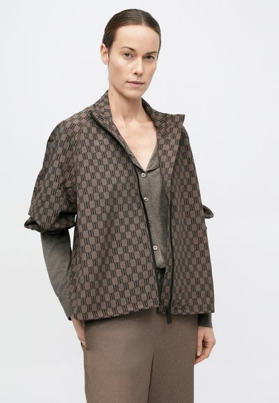 Jachetă maro cu model, prevăzută cu fermoar frontal, guler înalt și mâneci scurte. Purtați peste un top gri și asortată cu pantaloni bej.