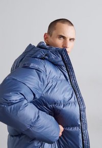 Gepolsterte blaue Pufferjacke mit hohem Kragen, Reißverschluss und strukturierter Oberfläche. Verfügt über eine taillierte Silhouette und verstellbare Kordeln.