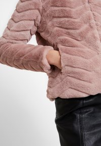 Osoba ubrana w miękką różową kurtkę z futra syntetycznego, z ręką w bocznej kieszeni, w połączeniu z czarnymi skórzanymi spodniami.