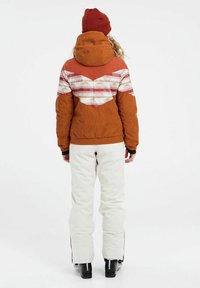 Oranje corduroy jacket met een patronen ontwerp op de achterkant, gecombineerd met witte broek en zwarte sneeuwlaarzen. Een warme gebreide muts maakt de outfit compleet.