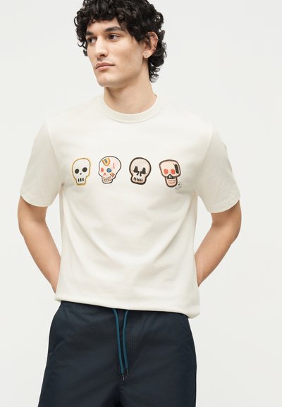 Νεαρός άντρας φορεί ένα λευκό μπλουζάκι με τέσσερα πολύχρωμα σχέδια κρανίων στο στήθος και σκούρο μπλε παντελόνι με κορδόνι.