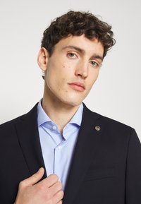 Giacca nera con una camicia a pois blu navy sotto. Presenta un emblema dorato sul rever e una texture liscia.