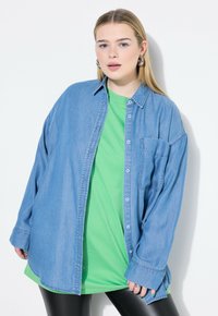 Camicia in denim blu chiaro con bottoni sopra una t-shirt verde, abbinata a leggins neri. Caratterizzata da una tasca sul petto e design ampio.